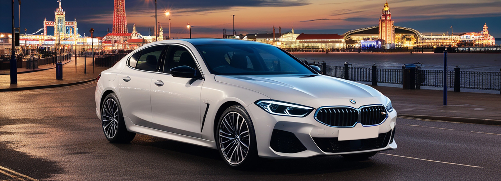 A white BMW Luxury Sports car parked on a seaside street with the iconic Blackpool Tower and illuminated buildings in the background at dusk